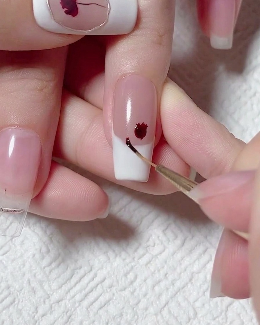 Gros plan dun pinceau liner appliquant un minuscule point de gel rouge foncé à côté dune fleur rouge plus grande sur un ongle French.