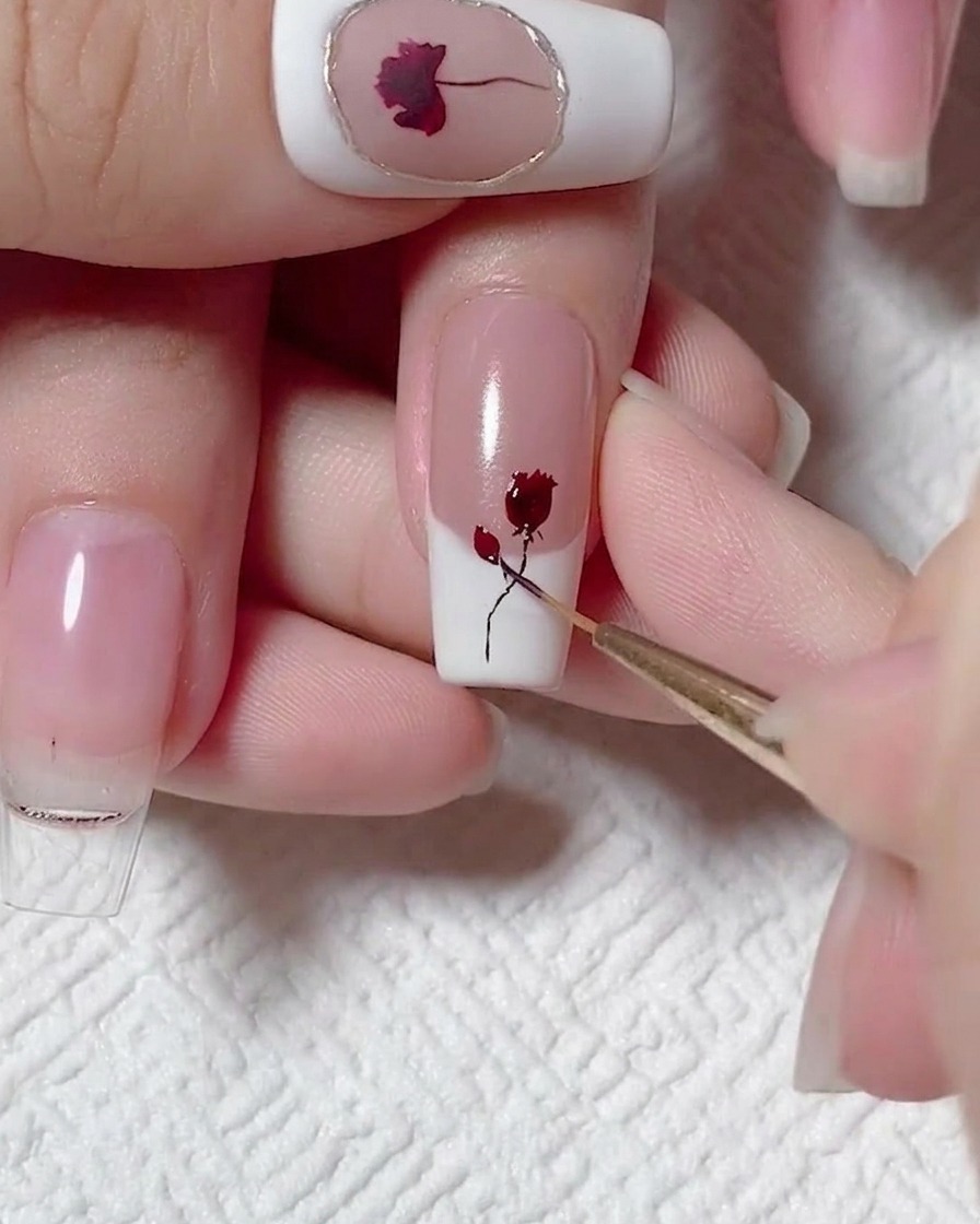Un pinceau liner ajoutant de minuscules feuilles sombres à une tige noire peinte et un design de rose rouge sur un ongle.