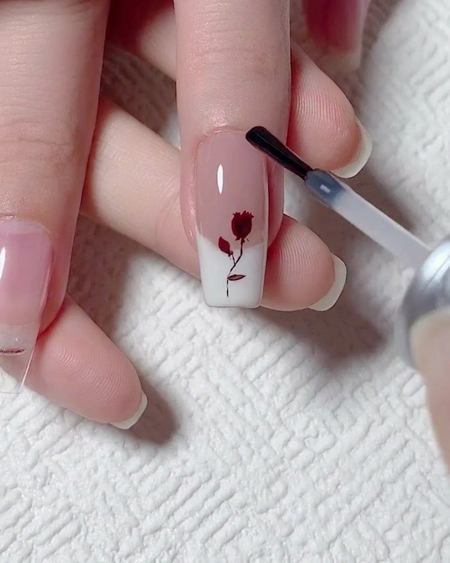 Application dune couche de top coat mat avec un pinceau sur un ongle avec une rose rouge et une French blanche.