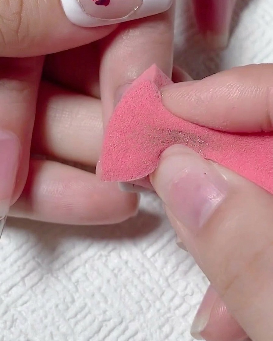 Une main utilisant une éponge à maquillage rose sèche pour essuyer lexcès de poudre chromée dorée sur un ongle mat.