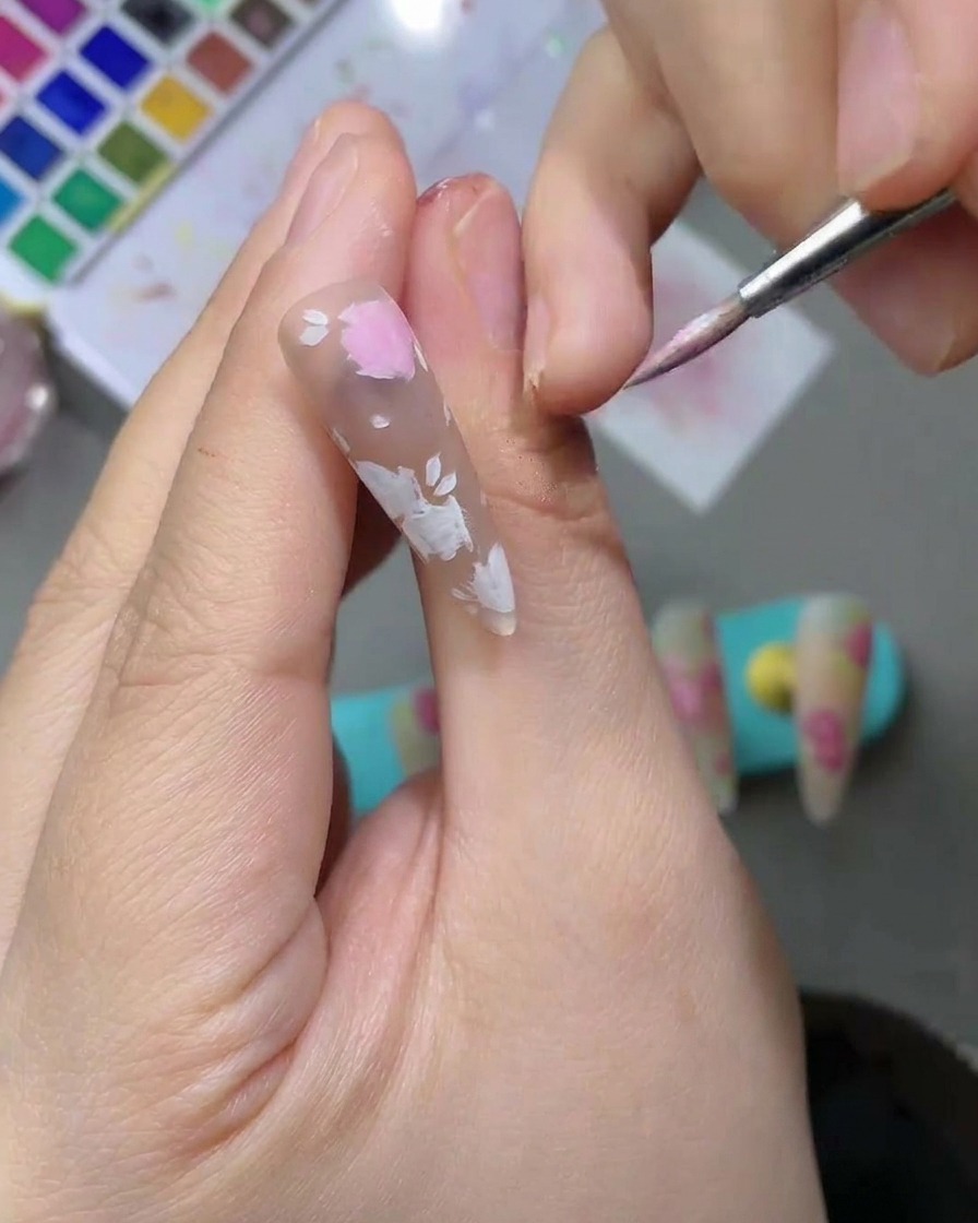 Gros plan sur un ongle avec des formes de pétales aquarelle rose clair délicates remplies à laide dun pinceau nail art à pointe fine.
