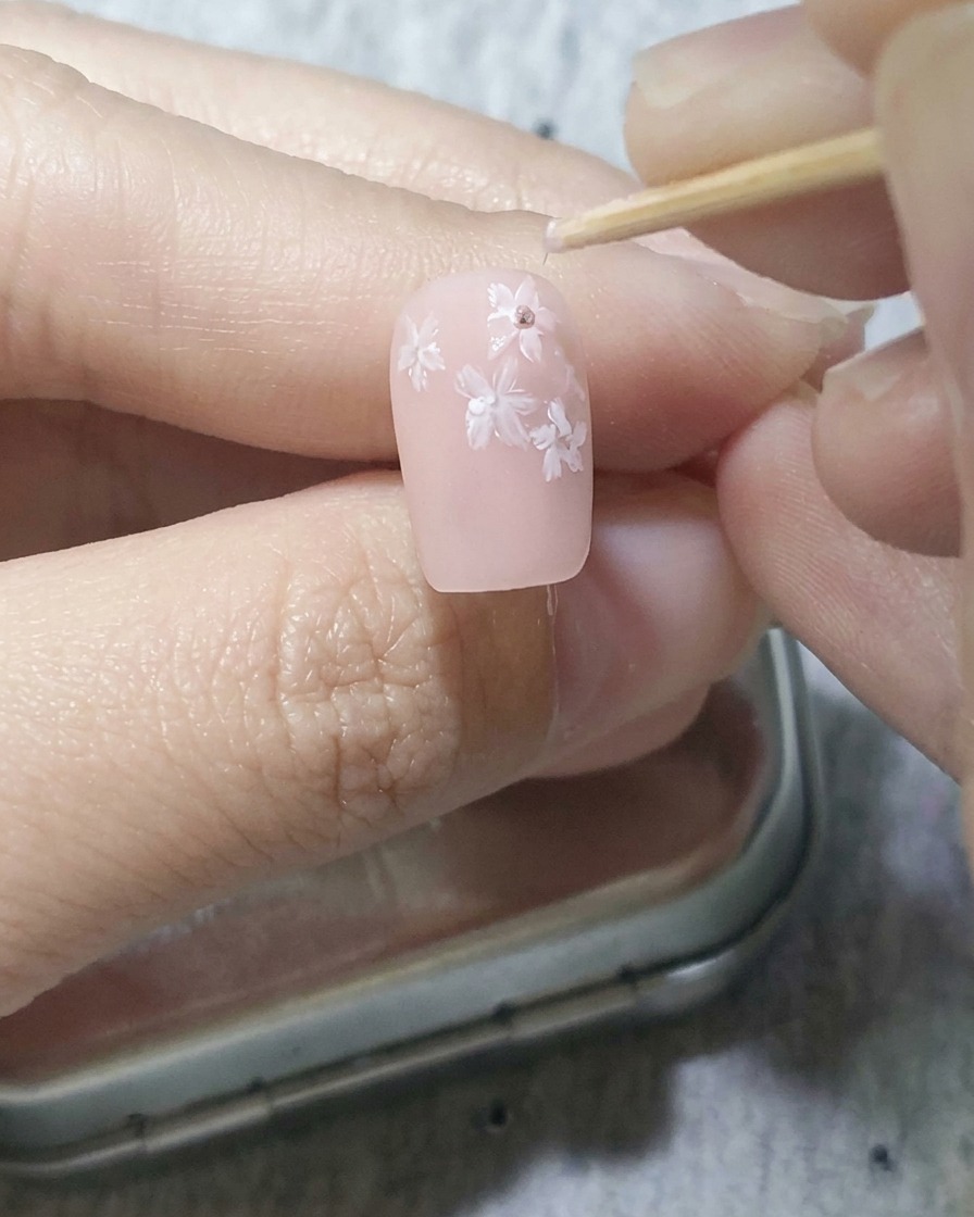 Un cure-dent plaçant doucement des points rose foncé au centre de fleurs blanches sur un ongle rose doux.