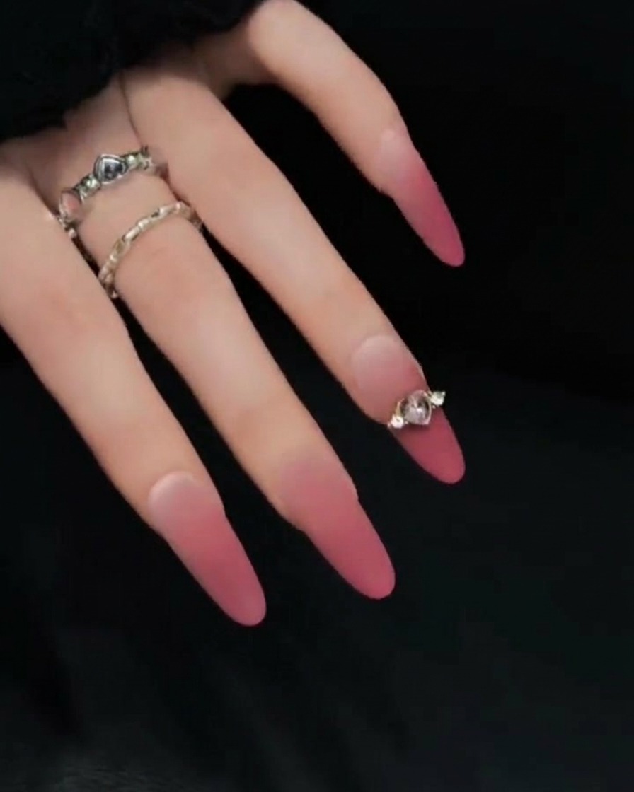 Gros plan dune main posée sur un fond sombre, montrant le profil latéral de longs faux ongles rose et nude pour souligner leur longueur et courbure.