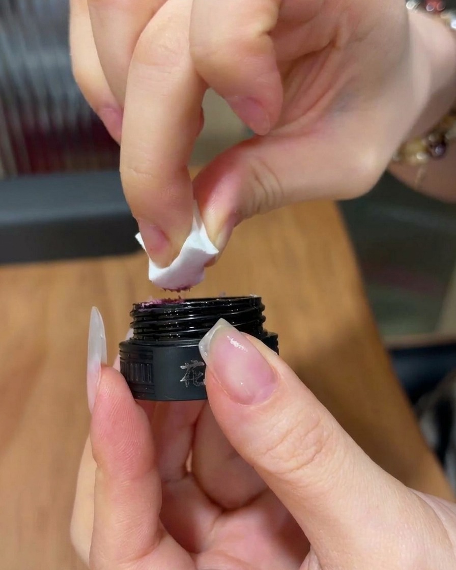 Gros plan sur une main tenant une éponge à maquillage triangulaire, prélevant une petite quantité de gel rose vif dans un pot cosmétique noir.