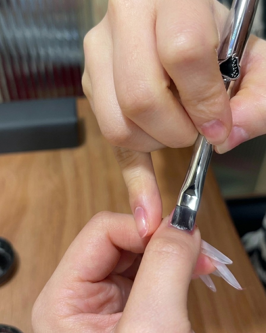 Un pinceau plat appliquant une fine couche de gel œil-de-chat scintillant sur un ongle présentant un dégradé rose.