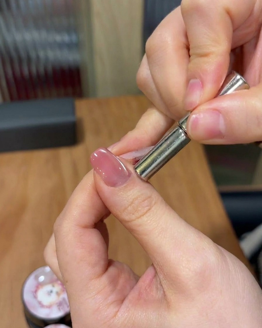 Une main tenant un aimant cylindrique au-dessus dun ongle, effectuant un mouvement pour manipuler les particules magnétiques en une bande diagonale scintillante.