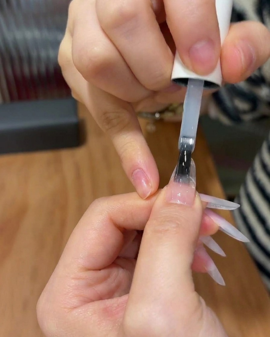 Un pinceau à ongles appliquant un top coat transparent et brillant sur un ongle présentant un design dégradé rose fini.