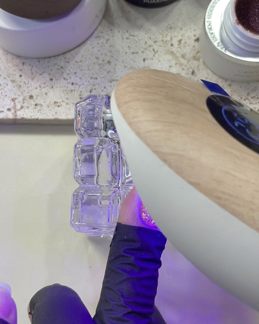 Une main positionnée sous une lampe UV/LED pendant la catalysation dun ongle avec des paillettes roses.