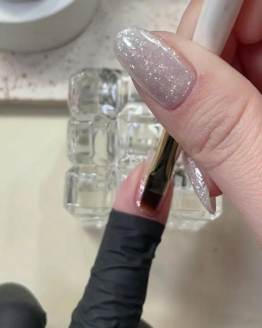 Un pinceau appliquant une fine couche transparente de gel de construction sur un ongle pailleté.