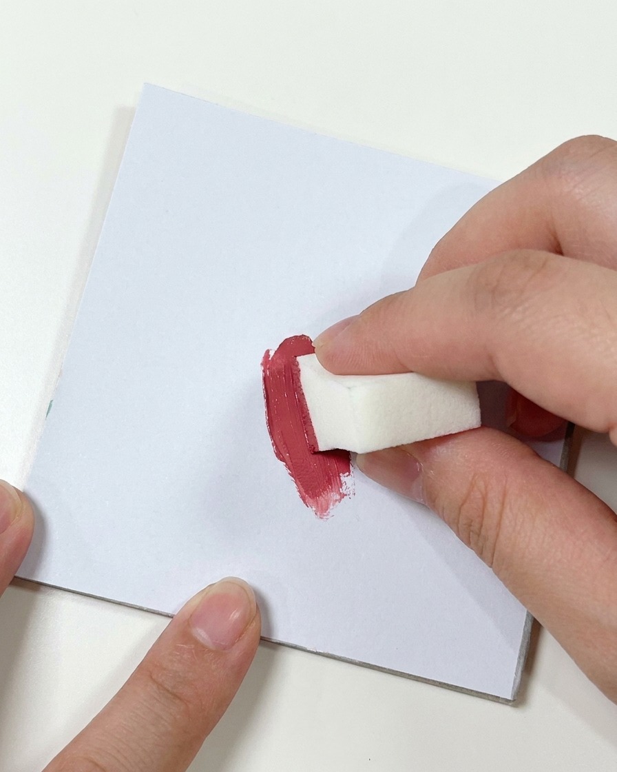 Une main pressant une petite éponge de maquillage blanche dans une touche de gel rose foncé sur une palette blanche plane.