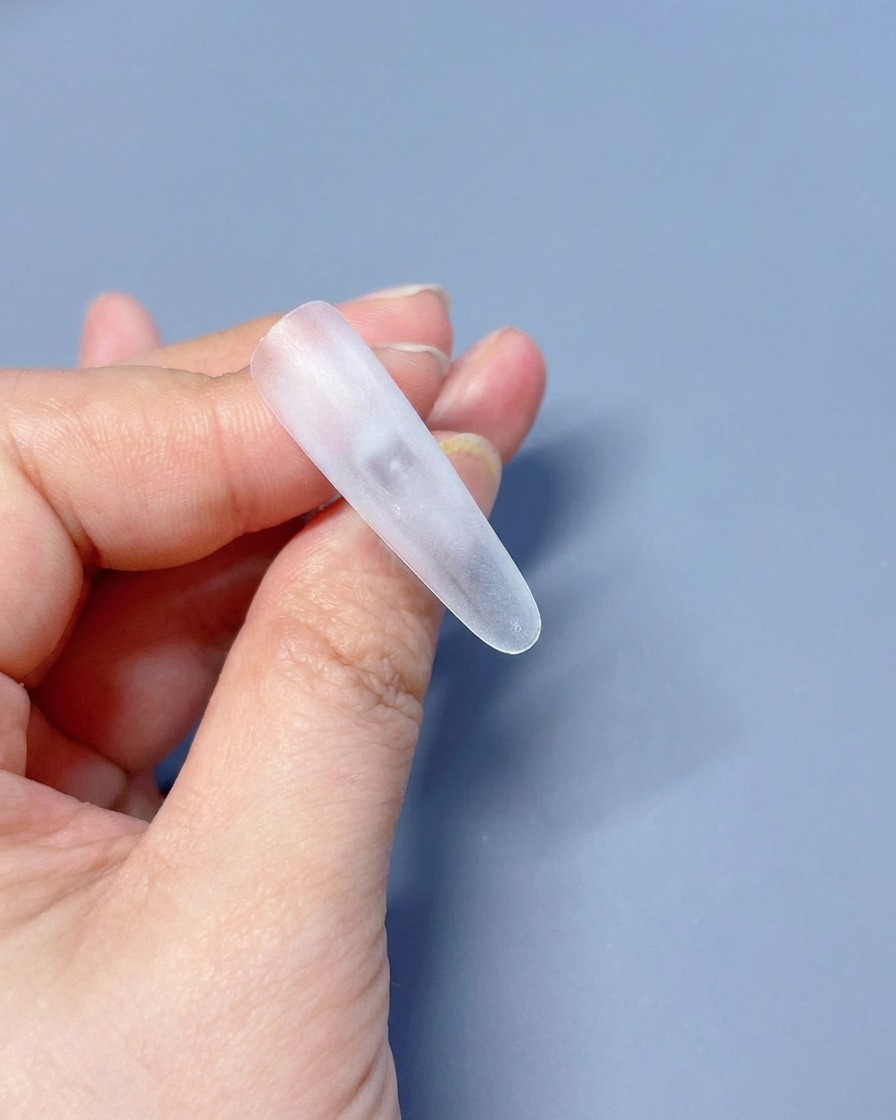 Gros plan sur une capsule press-on transparente tenue entre les doigts, prête pour lapplication de la base.