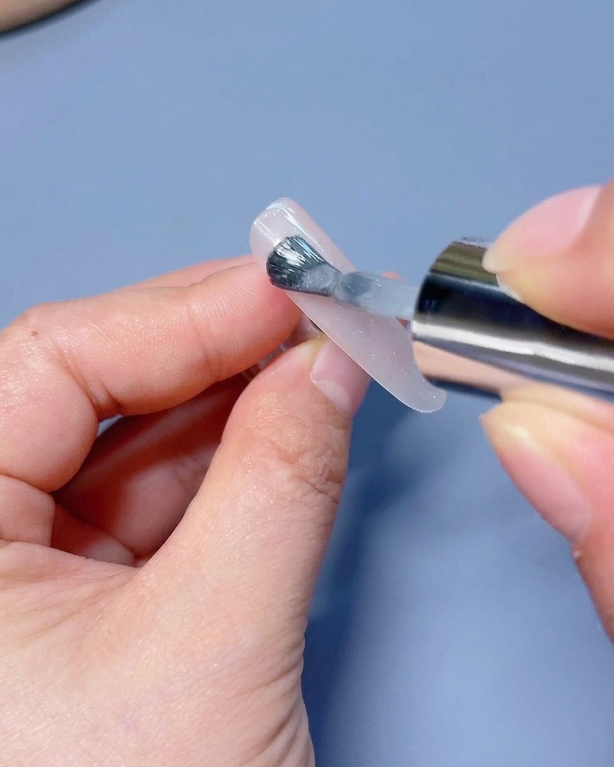Un pinceau appliquant une couche de vernis gel œil-de-chat blanc scintillant sur une capsule.