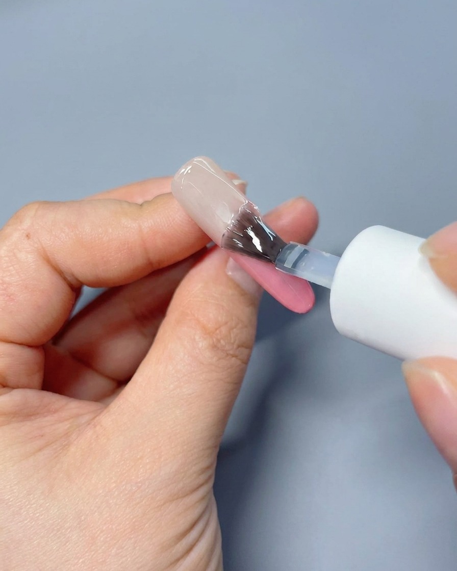 Un pinceau appliquant un top coat brillant sur un ongle au dégradé rose doux.