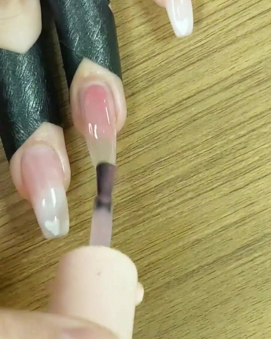 Un pinceau appliquant du vernis gel nude transparent sur toute la surface de longle.