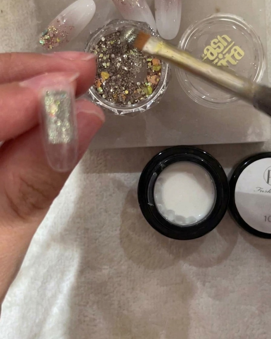 Un pinceau dépose des paillettes argentées et irisées sur la moitié supérieure dune capsule à ongle.
