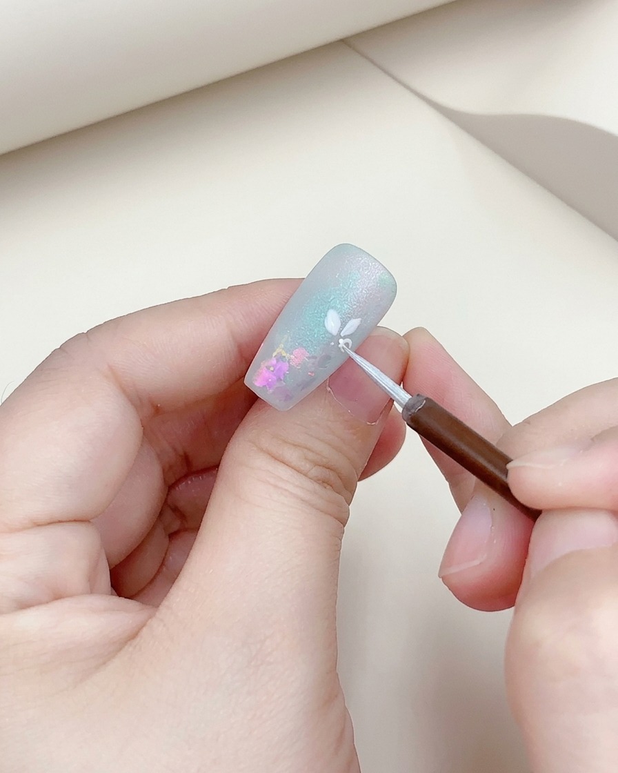Gros plan dun pinceau fin ajoutant un contour argenté à une fleur peinte en blanc sur un ongle.