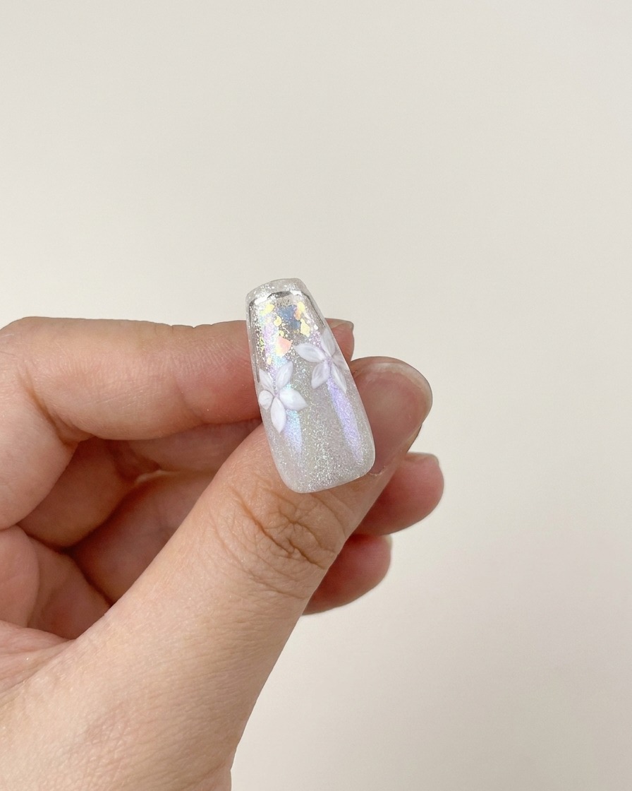 Un pinceau appliquant une couche de top coat brillant sur un design dongle fini avec des fleurs blanches et des flocons argentés.