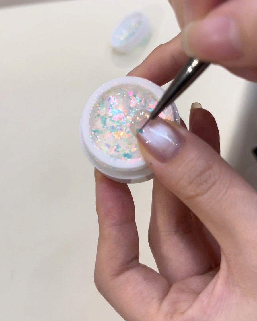 Une pince déposant délicatement des flocons irisés bleus sur un ongle enduit de gel de construction humide.