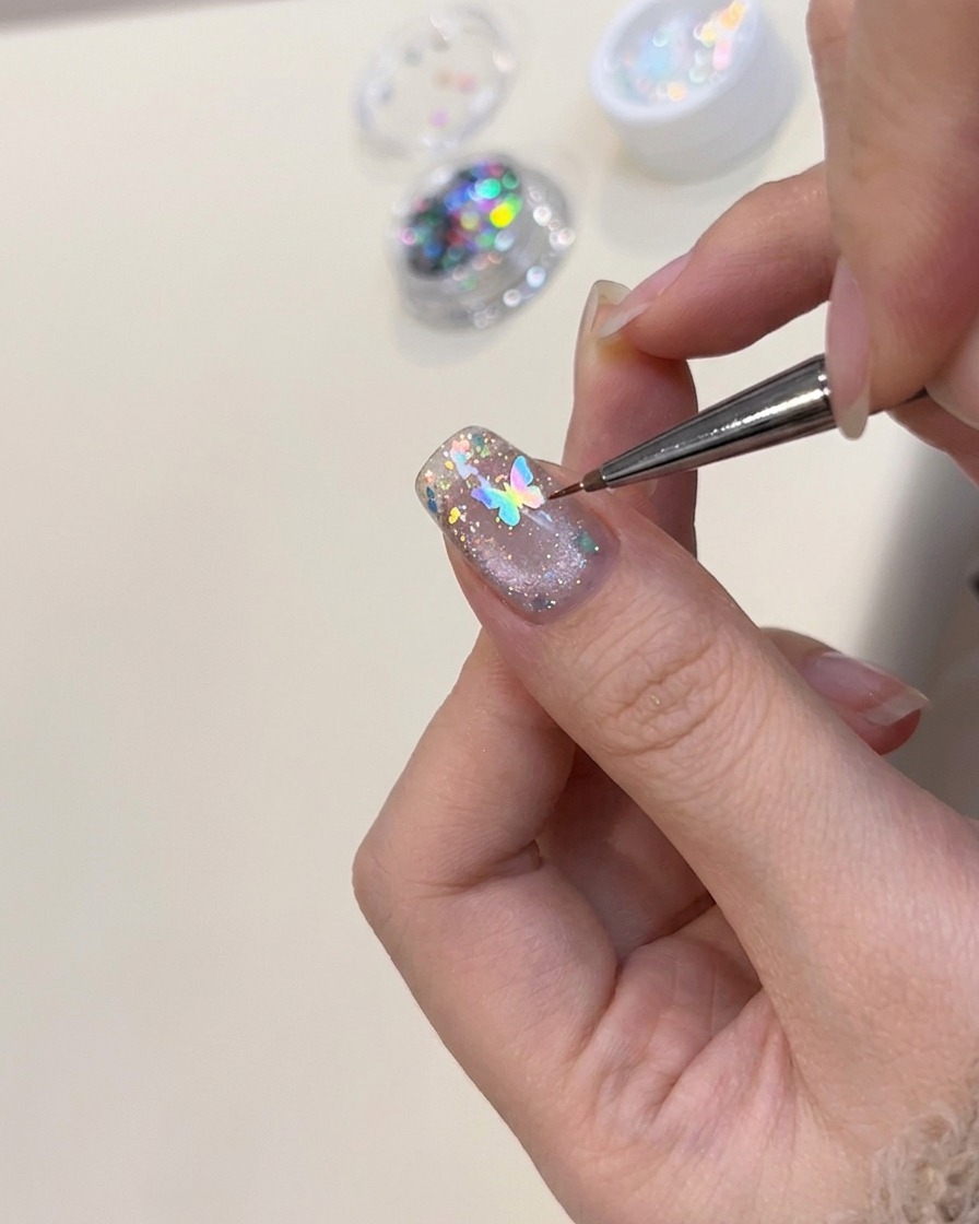 Une pince positionnant un petit charme papillon holographique sur un ongle décoré.