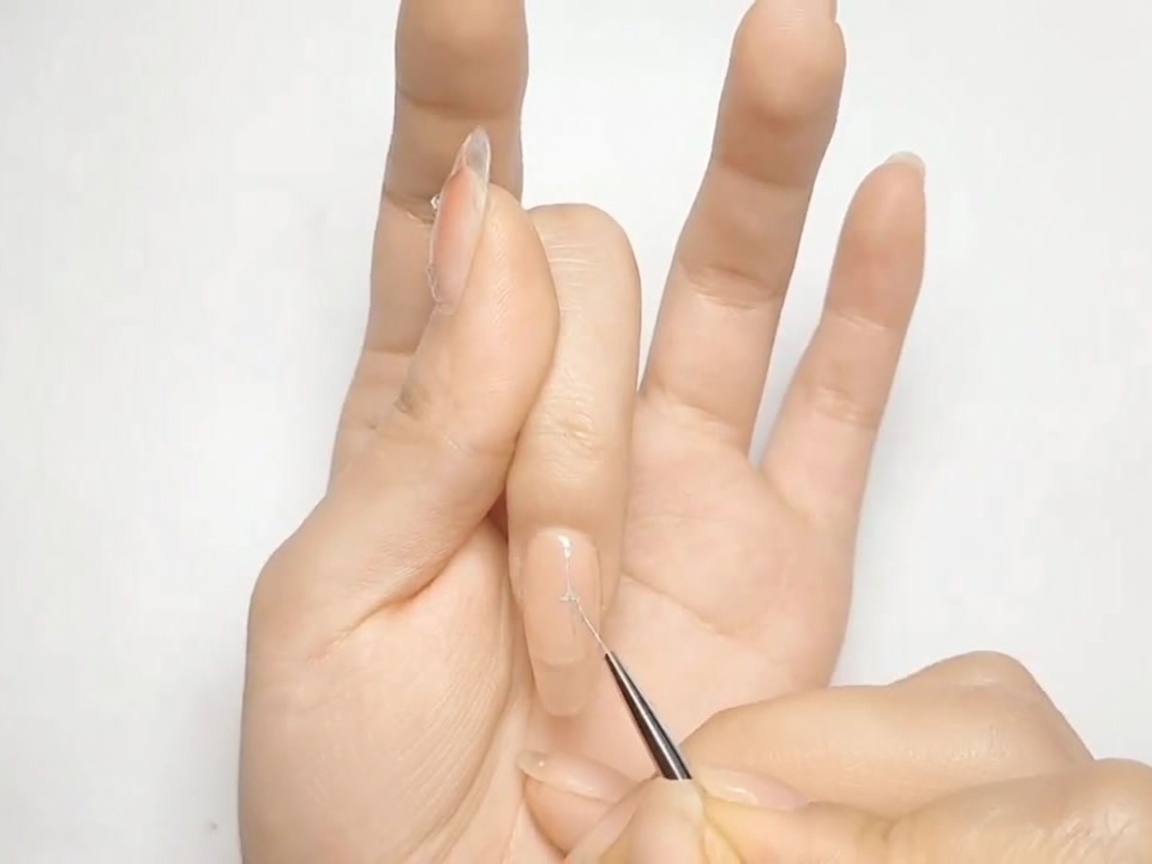 Un pinceau liner fin dessinant une croix argentée sur un ongle peint en nude.