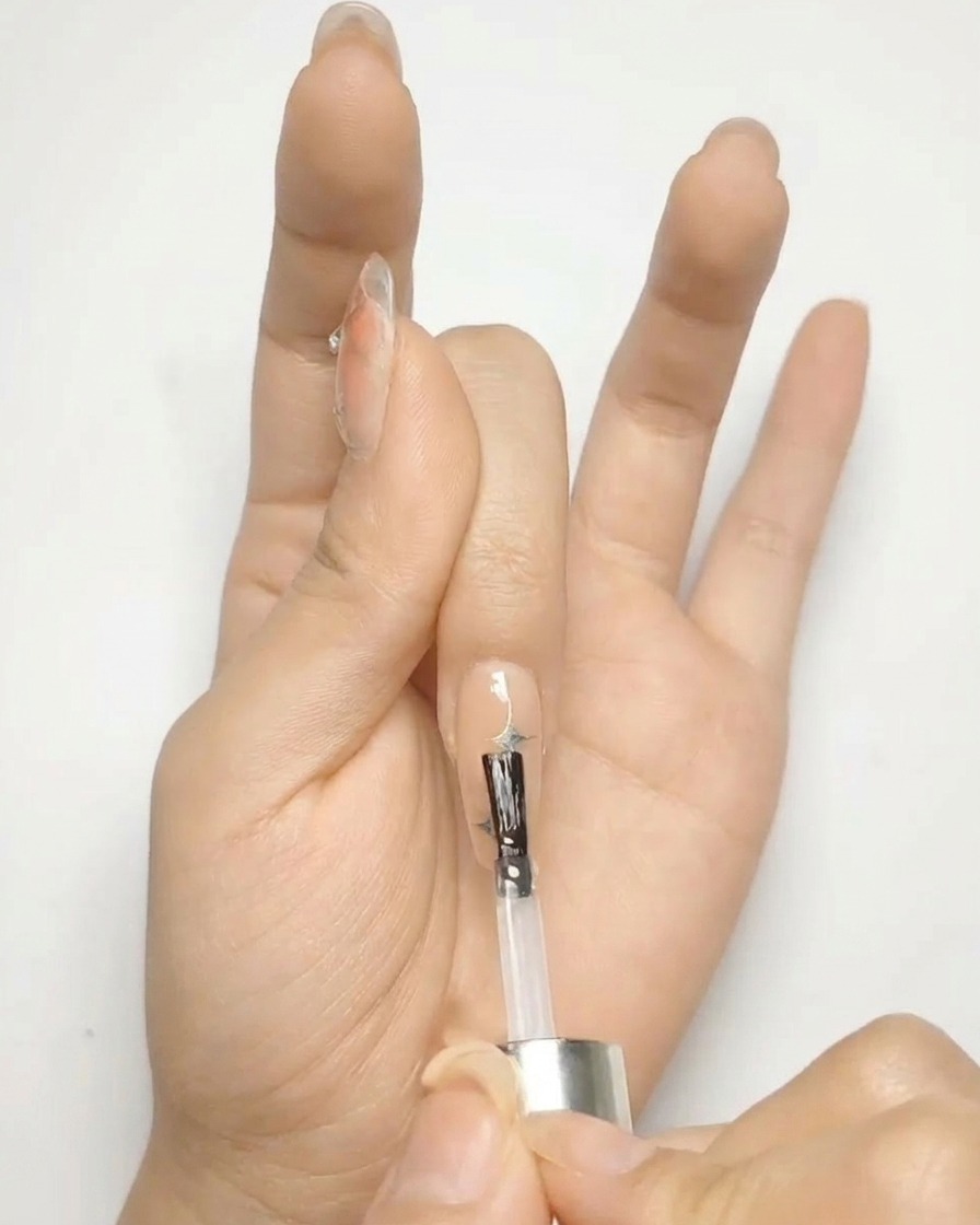 Un pinceau appliquant un top coat transparent brillant sur un ongle nude décoré détoiles argentées.