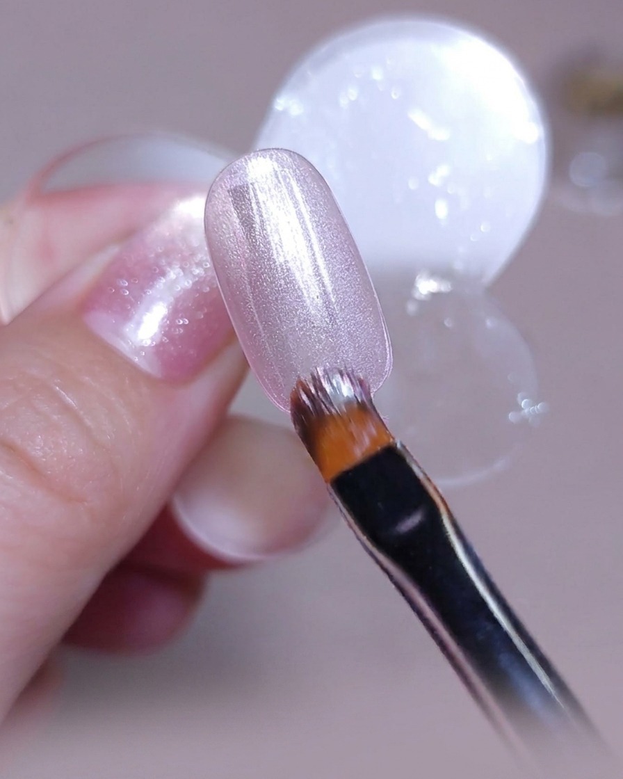 Un pinceau appliquant une couche uniforme de vernis gel cat-eye rose argenté sur une capsule dongle transparente.