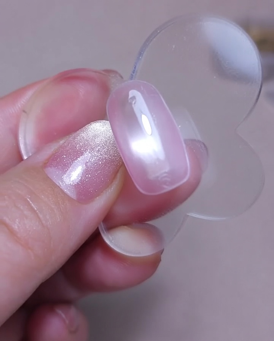 Vue rapprochée dun aimant tenu au-dessus dun ongle avec du gel cat-eye rose encore humide pour créer un effet scintillant.