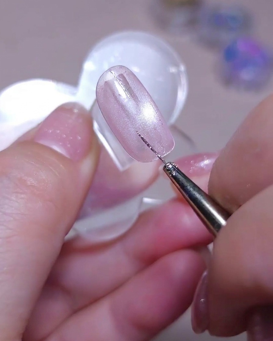 Un pinceau liner fin dessinant une deuxième ligne verticale droite de paillettes argentées à côté dune ligne existante sur un ongle cat-eye rose scintillant.