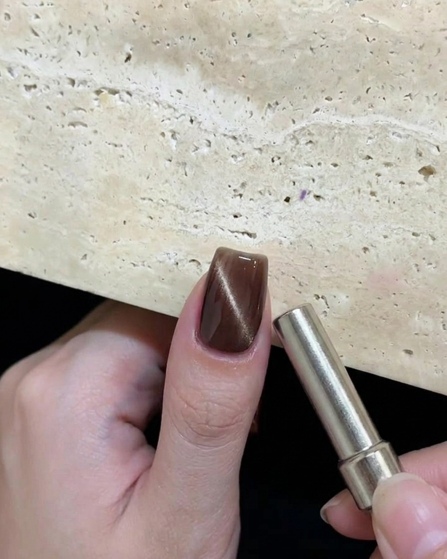 Une main tenant un aimant cylindrique près dun ongle avec du gel œil de chat marron humide pour créer un motif circulaire réfléchissant.
