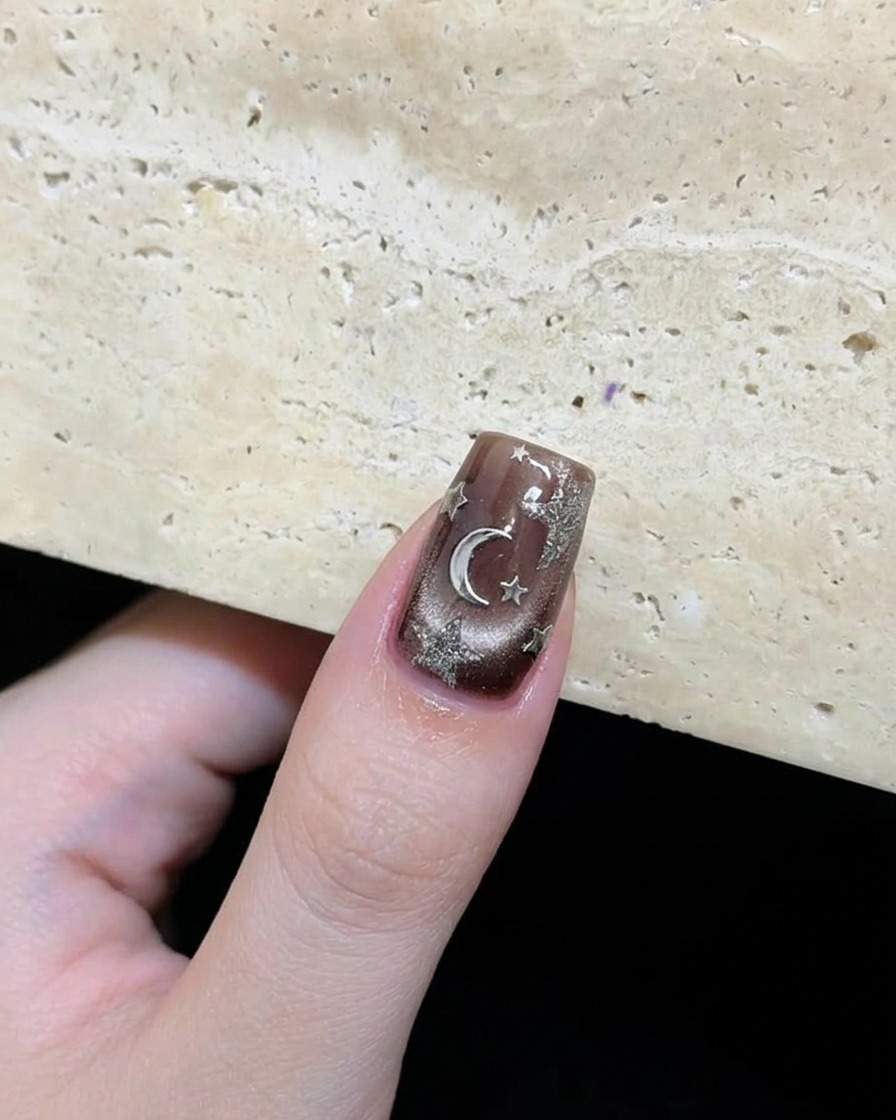 Gros plan dun ongle marron foncé terminé avec des étoiles argentées et un accent en forme de lune disposés de manière artistique.