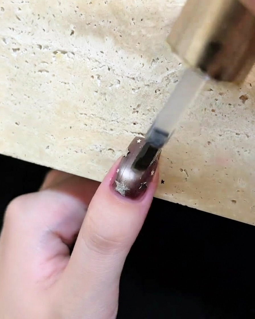 Un pinceau appliquant un top coat transparent et brillant sur un ongle marron foncé décoré détoiles et de lunes argentées.