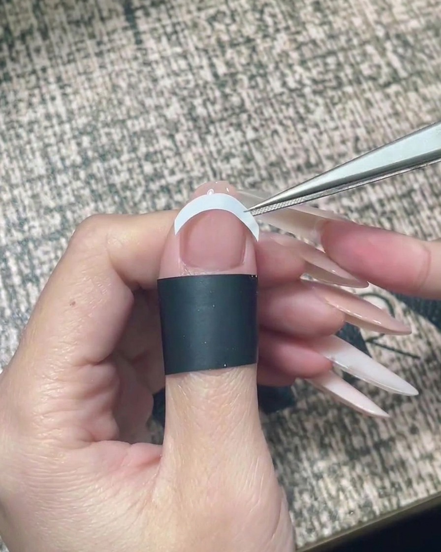 Une pince à épiler plaçant un autocollant guide blanc courbé pour French manucure horizontalement au milieu dun ongle.
