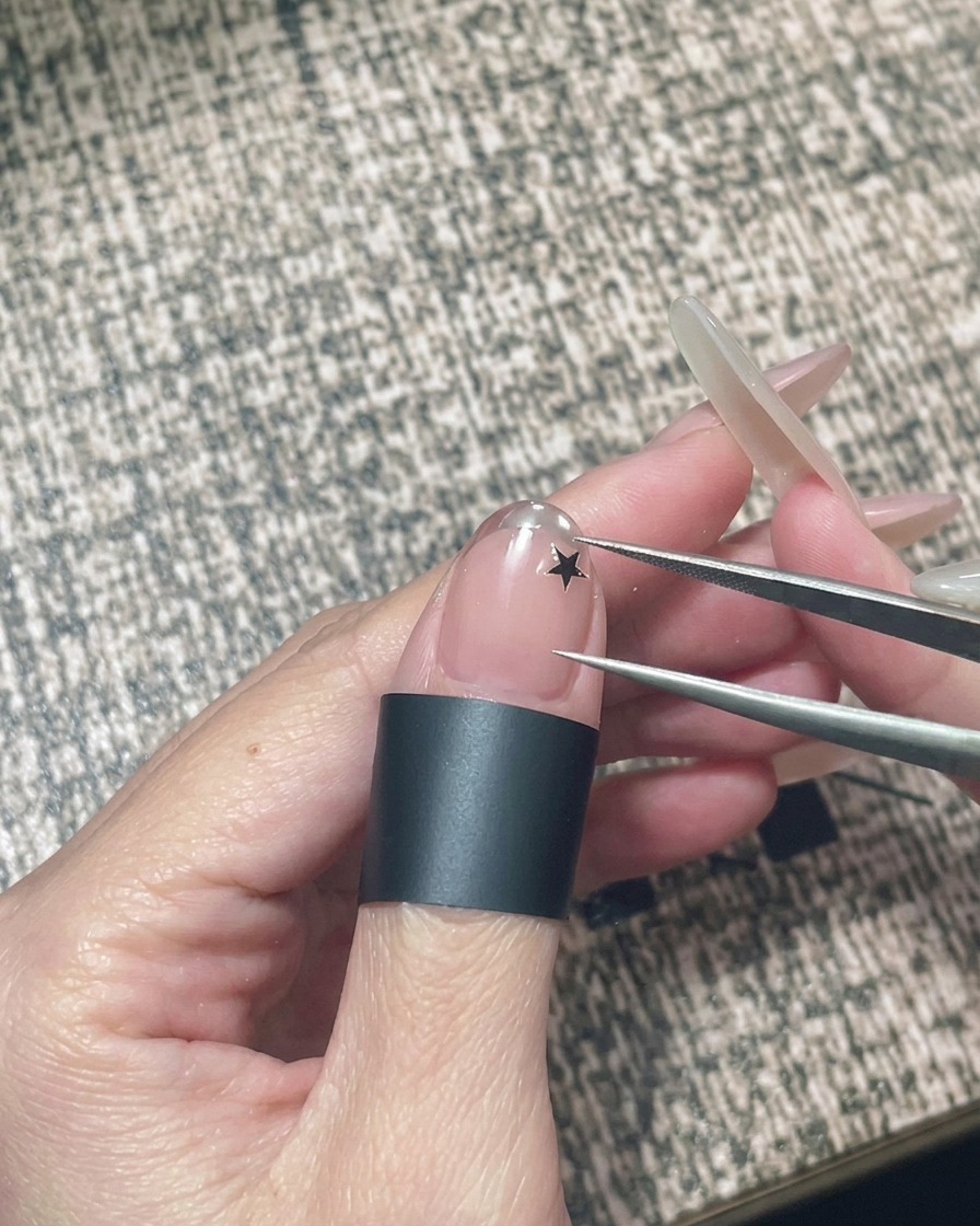 Une main tenant une pince à épiler pour placer un petit autocollant étoile noire sur un ongle en gel nude.
