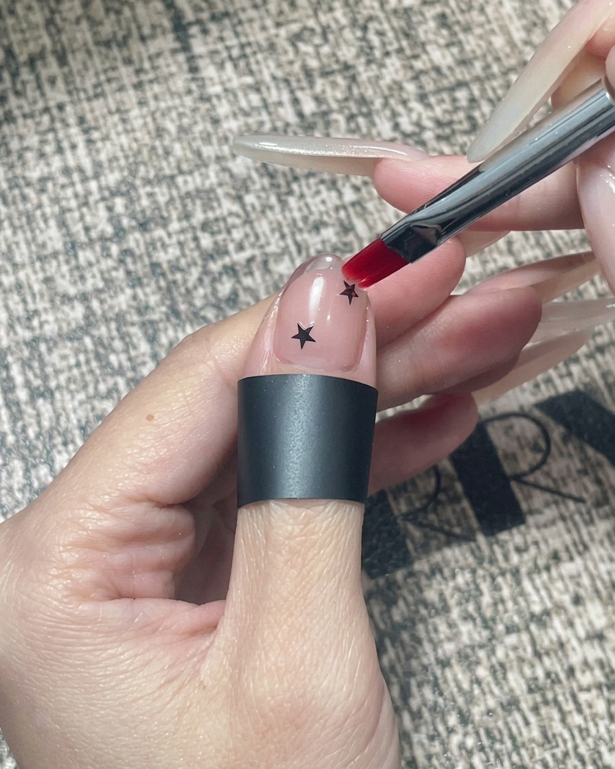 Un pinceau à gel carré appliquant un gel encapsulant transparent sur un ongle décoré détoiles noires et dun bout chromé.