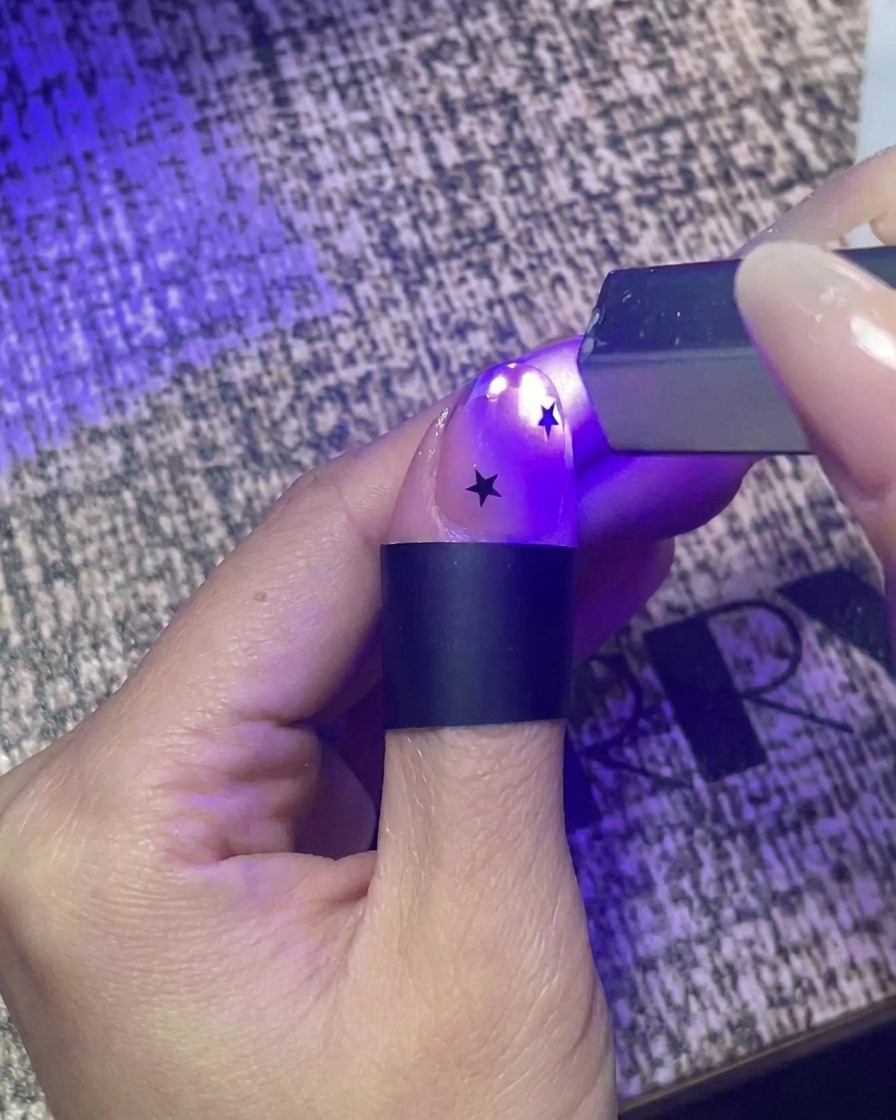 Gros plan dune main utilisant une petite lampe de poche UV portative pour catalyser un ongle décoré dautocollants étoiles noires.