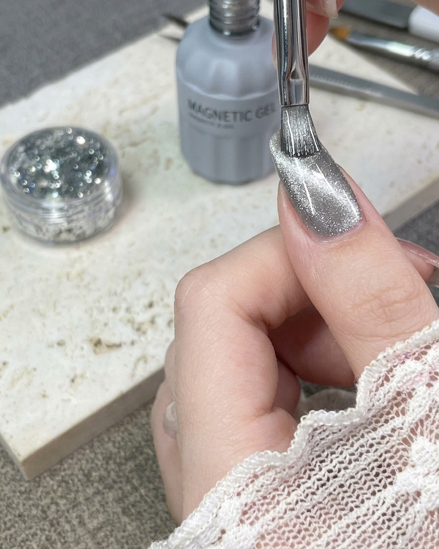 Un pinceau appliquant une couche de vernis semi-permanent argenté œil-de-chat sur un ongle naturel.