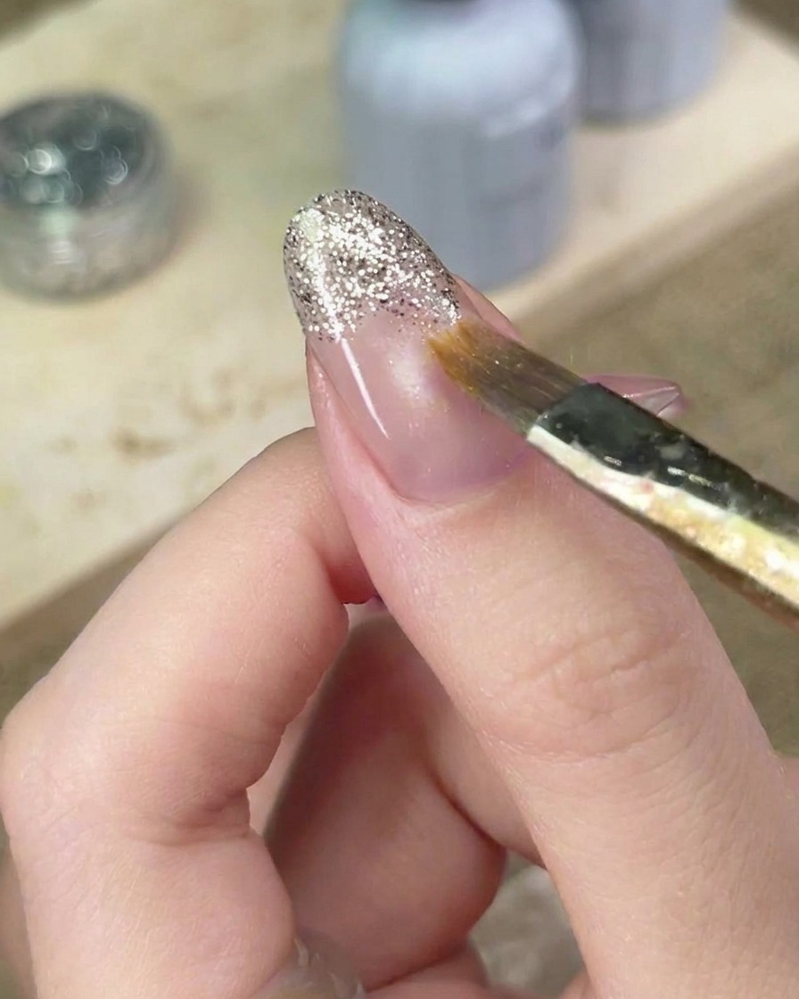 Un pinceau de nail art estompant soigneusement les bords du gel pailleté argenté sur longle pour parfaire le dégradé.