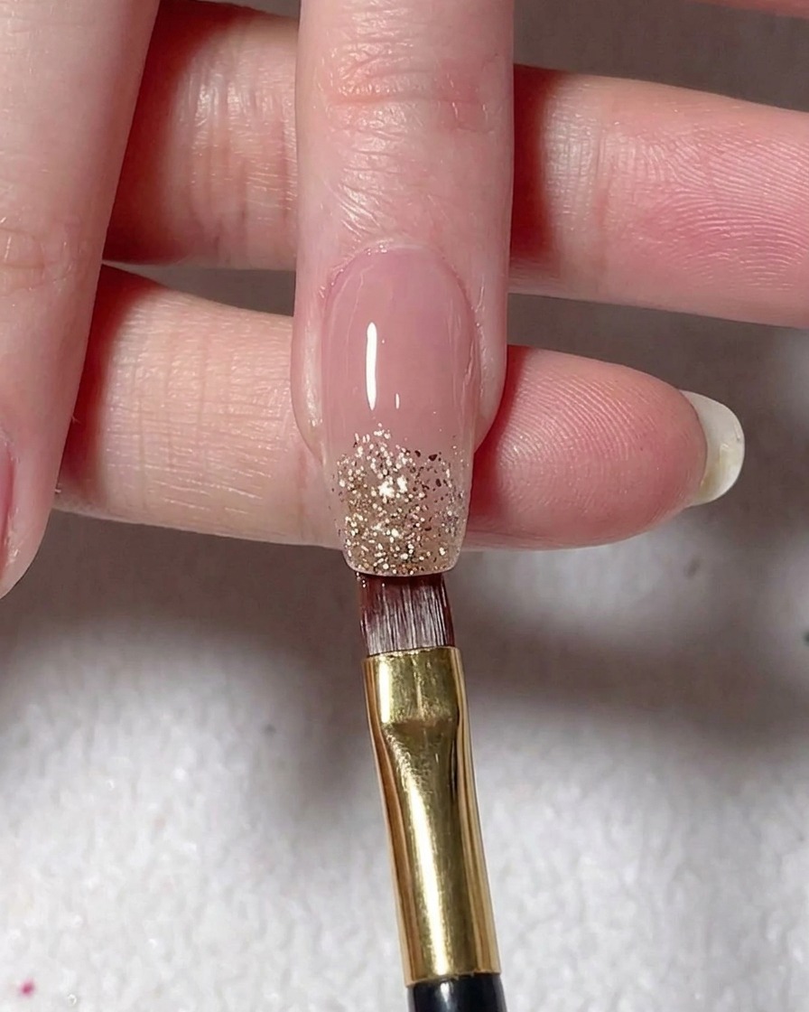 Un pinceau plat déposant du gel à paillettes argentées sur le bout dun ongle peint en nude.