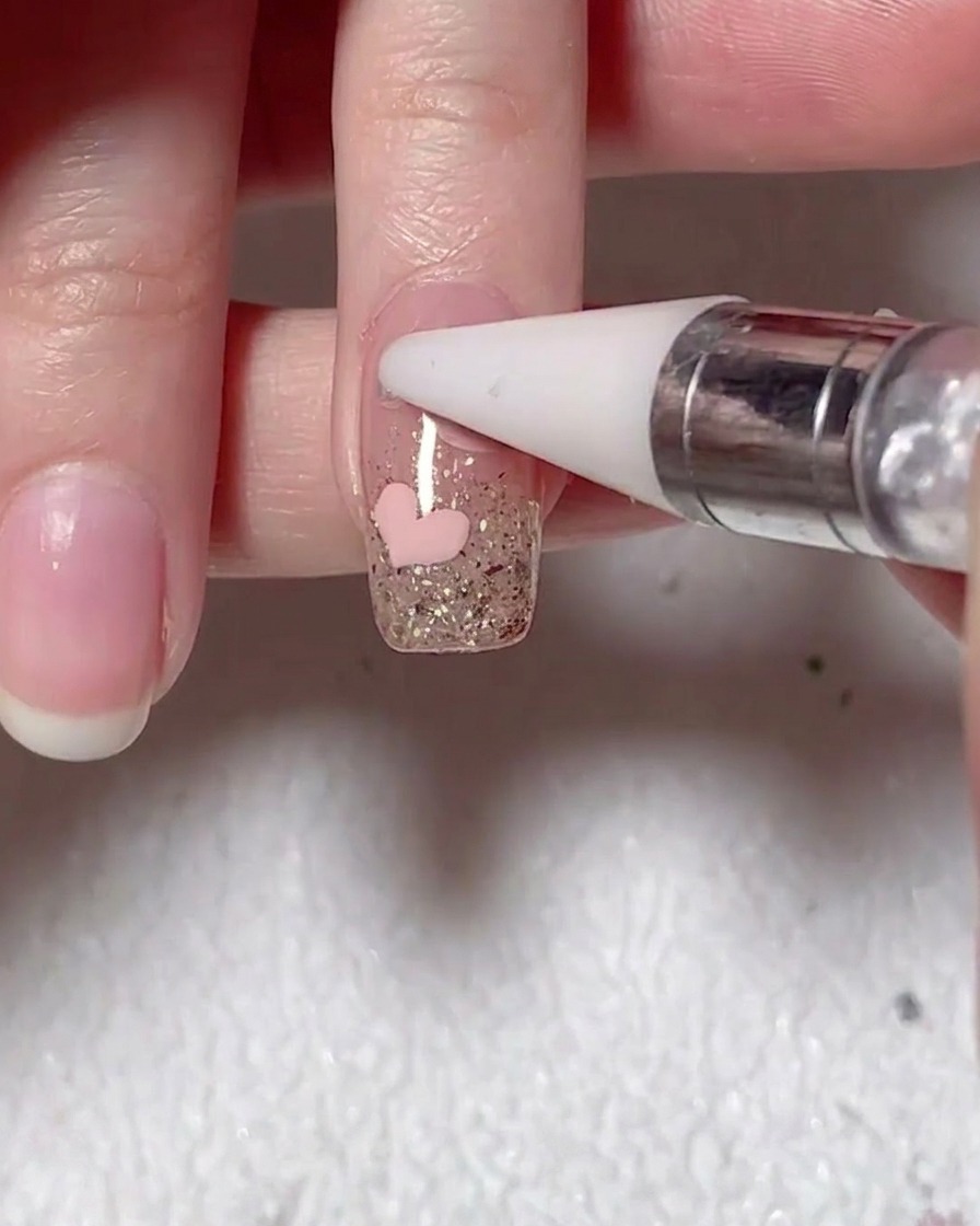 Un outil à embout de cire plaçant une petite perle sur la surface de longle à côté du cœur rose.
