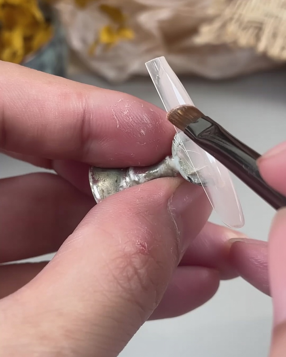 Un pinceau à ongles appliquant une couche de base de gel nude laiteux sur une capsule transparente tenue par des doigts.