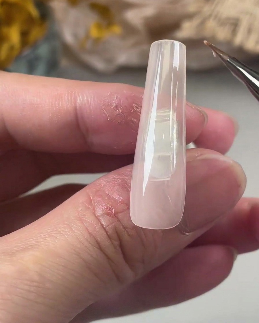 Un pinceau de détail fin appliquant une couche lisse de gel de base transparent sur une longue capsule carrée de couleur nude.