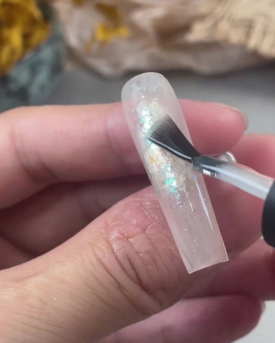 Un pinceau à ongles appliquant une couche épaisse de gel de renforcement transparent sur un ongle à paillettes irisées.