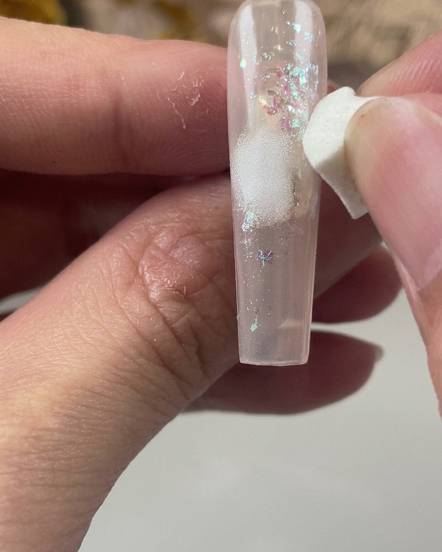 Une éponge cosmétique blanche tamponnant doucement une tache de peinture blanche au centre dun ongle encapsulé de paillettes.