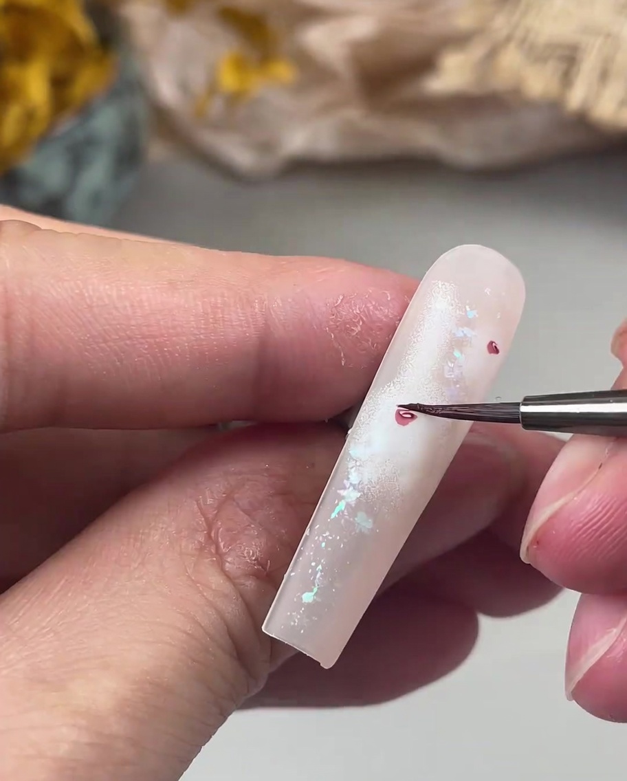 Un pinceau fin pour nail art peignant de délicats petits pétales rose foncé autour dun cœur de rose central sur un ongle.
