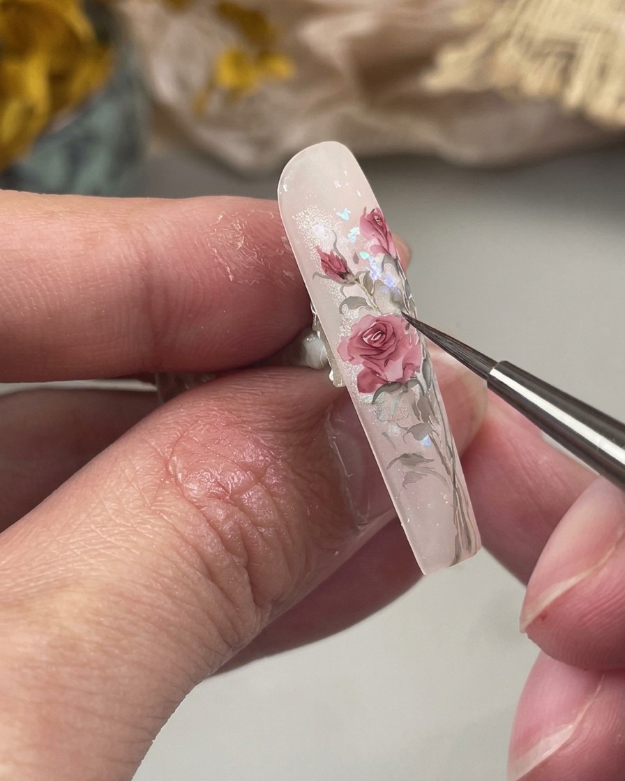 Une photo en gros plan montrant un pinceau fin peignant de petites feuilles vertes effilées à la base dun motif floral couleur rose sur longle.