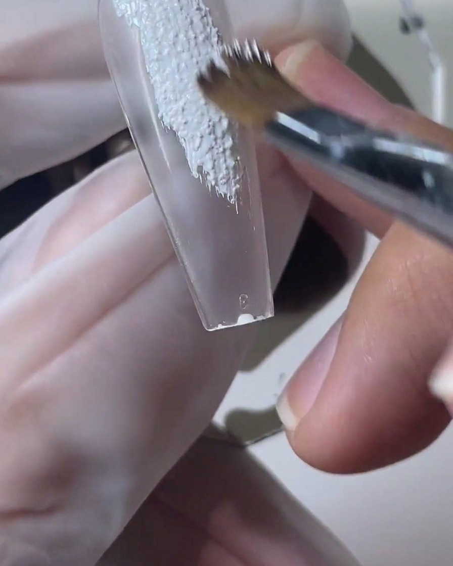 Un pinceau à ongles applique du gel sable texturé blanc sur une capsule transparente.