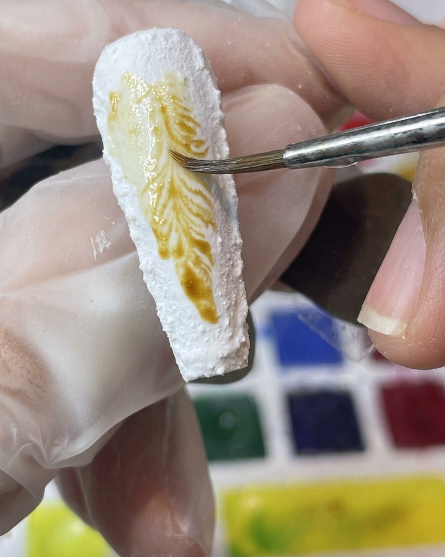 Un pinceau applique des pigments jaunes et marron sur une surface dongle blanche texturée.