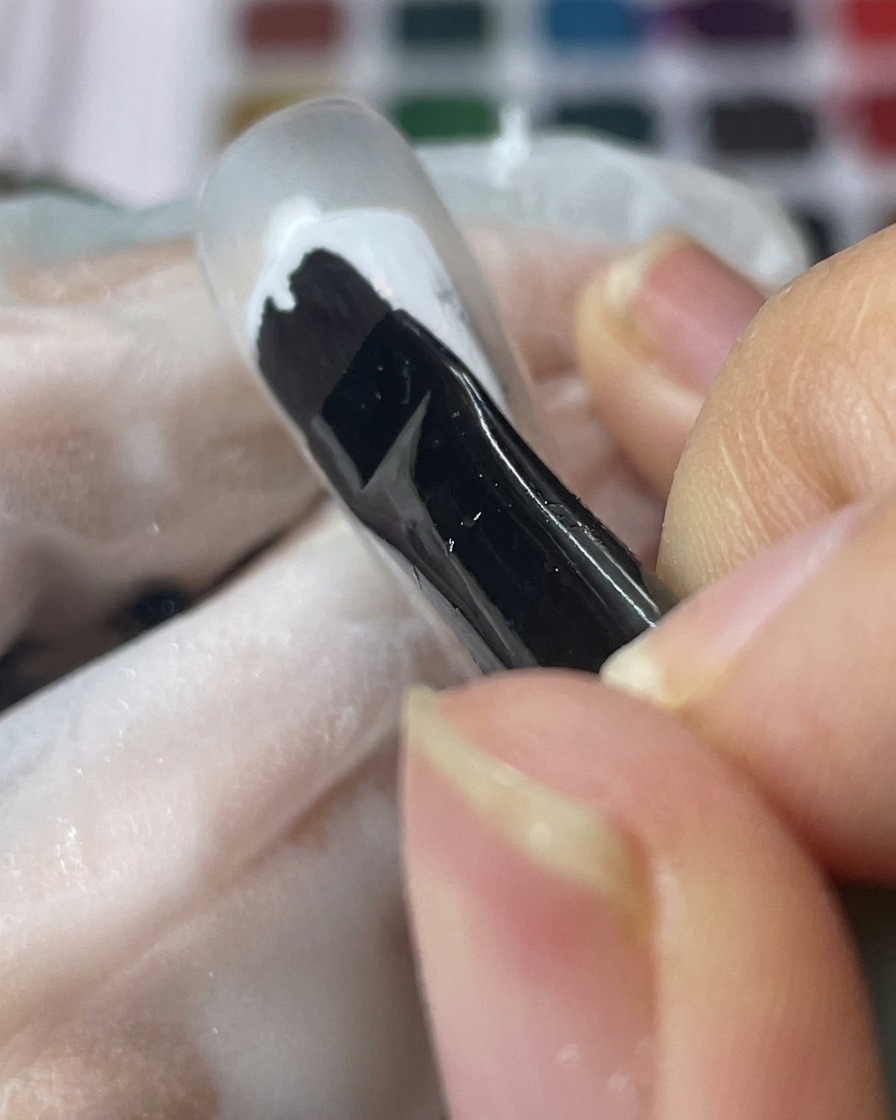 Un pinceau pour gel noir appliquant une couche lisse de gel blanc sur une capsule transparente.