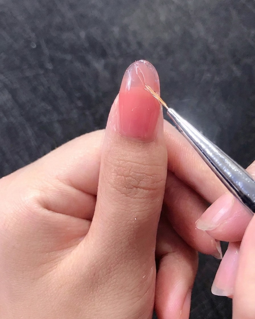 Gros plan sur un pinceau à liner dessinant de fines lignes dorées sur un ongle en gel rose.