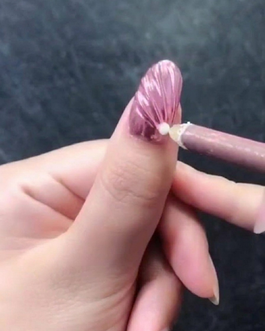 Gros plan dune main avec un ongle texturé rose métallique, montrant un stylo à cire plaçant une perle blanche unique près de la cuticule.